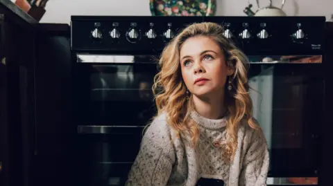 Bryony Coles Saoirse-Monica Jackson has long blonde curly hair. She is facing the camera but looking up. She has a knitted jumper on and is sitting in front of a cooker.