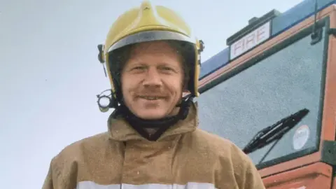 Steve Paige An old photo of Steve who is wearing a yellow helmet and is in light brown firefighting gear