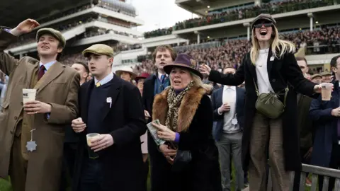 PA A group of five people screaming and cheering as the horses pass them on the racecourse. The grandstands behind them are packed full of people.