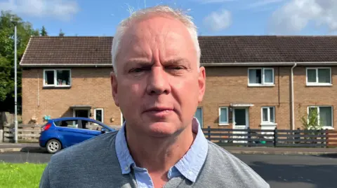 Dermot Nicholl is standing in a residential street. He is wearing a grey jumper and a blue shirt. A number of houses are visible in the background with a few cars parked outside them.