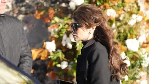 Andrew Matthews/PA Media Cheryl Tweedy in black mourning clothing and sunglasses near a vehicle with a man in a black coat visible next to it, and flora behind them.