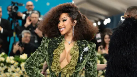 Singer Cardi B wears a velvet green suit and fluffed hair on the Met Gala blue carpet.