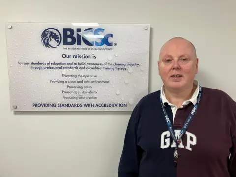 James Grant/ BBC A man in a navy long sleeve polo shirt stands in front of the British Institute of Cleaning Science logo