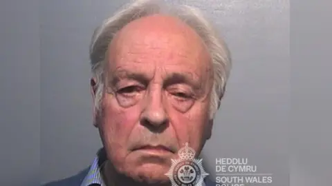 A mugshot of Anthony Pierce who has white hair and is wearing a striped blue shirt and blue sweatshirt and is looking directly at the camera with a neutral expression