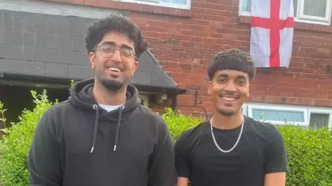 Two men stand outside a house smiling. An England flag is hanging out of one of the top windows of the house