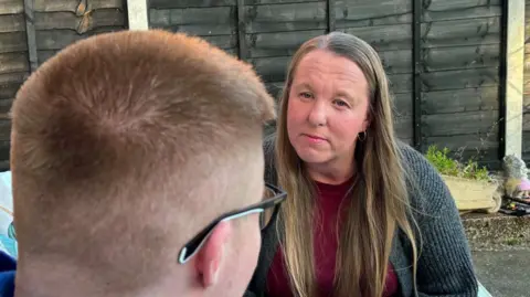 Family handout A woman facing towards her son. We see the back of his head and he his wearing glasses. They are both sitting outside in front of a garden fence.