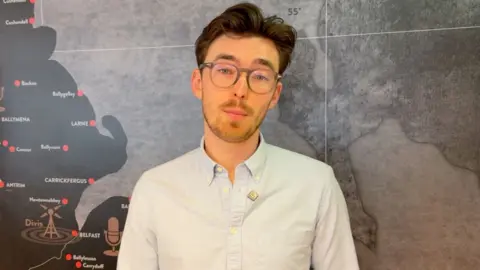 Patrick Fee in a blue shirt stood against a map of NI