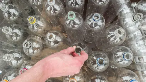 PA Media A hand touching the top of a clear bottle in a tray of clear bottles. 