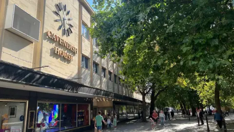 Cavendish House, a large department store building on Cheltenham's Promenade, glows in the sun. Shoppers are walking beneath leafy trees in the shade. There is a large clock on the front of the building.