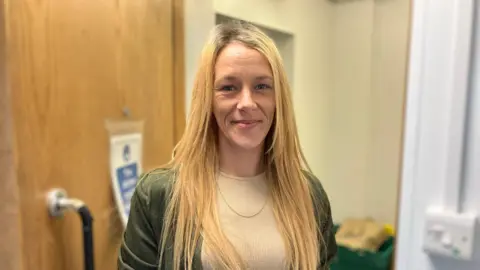 A woman with straight blonde hair in a beige dress with a green jacket on, smiles at the camera.