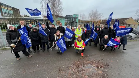 Stuart Woodward/BBC Group of people in winter clothing holding union flags outside a school fence. The school building - mainly glass and green cladding - is behind them.