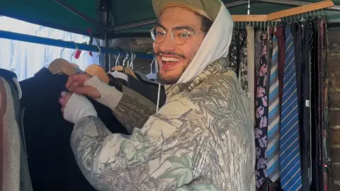 Khaled Ainsworth, who has a beard, is smiling at the camera. He is wearing glasses, a hooded sweatshirt, and a camouflage jacket. He is standing under a market-style canopy and browsing a rack of jackets and shirts on wooden hangers.