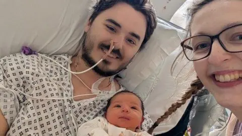 Handout Sam, Emma and Sophie King are pictured together in hospital. Sam is lying in bed in a hospital gown with various tubes going into his body. Emma is leaning into shot from the right and smiling at the camera. Between them, baby Sophie is held in Sam's arm.