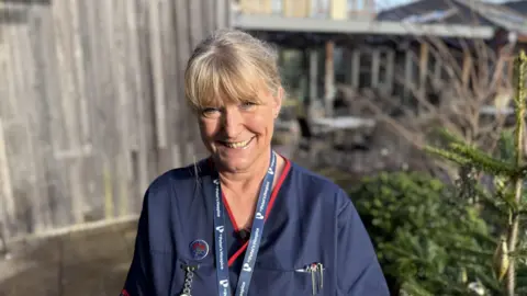 Juliette Hughes standing outside St Peter's Hospice in Bristol. She is wearing a dark blue healthcare tunic and is smiling.