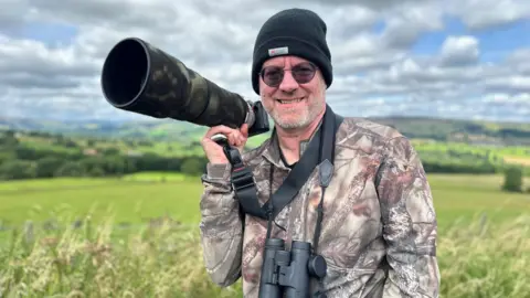 A man in a camouflage jacket with black binoculars hanging from his neck and a large camera being held in his hand smiles at the camera.