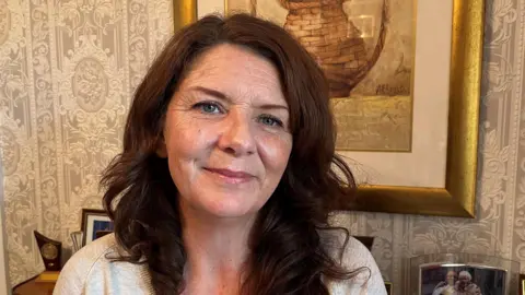 Rachel Austin, who has dark brown hair, sitting in a living room looking into the camera, with a picture behind her.