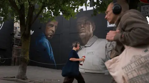Reuters People walk past a huge street mural which is a painting of Noel and Liam Gallagher. The oasis and adidas logos are also featured on the mural.