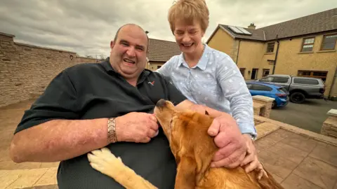 Marshall and his wife pet their dog, which looks like a Golden Labrador. It is jumped up on Marshall and has it's chin on his stomach. 