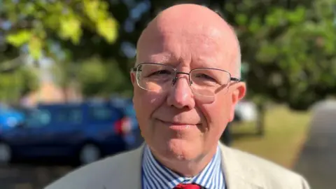 Dr Tim Allison standing outside. There are trees and a blue car in the background