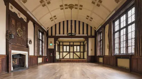 Historic England A large, empty hall. It has wooden flooring and the walls feature dark wooden panelling. To the left is a fireplace and to the right are two large windows.