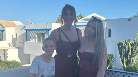 Leah Matthews A woman with brown hair in a bun, wearing a maroon jumpsuit with a black and gold belt and black cross-body bag. She has one arm around a young blonde boy in a red and white football shirt, and the other arm around a teenage girl with long blonde hair, wearing a maroon co-ord outfit and sunglasses on her head. The trio are stood on a white balcony with white-wash buildings and greenery behind them. They are all smiling and looking at the camera.