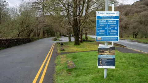 A car park is pictured with road markings, trees and grass by it. A sign listing the parking rules and fees is on a pole.