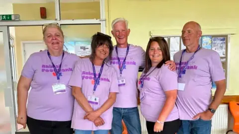 Animal Foodbank UK Five people are standing in a row, all wearing lilac-coloured T-shirts, emblazoned with the Animal Foodbank UK logo. They also have identity lanyards on and they are all smiling.