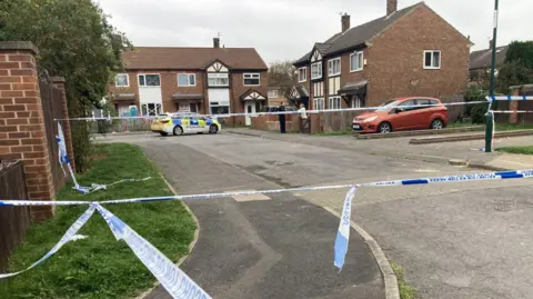  a small cul-de-sac consisting of a cluster of red-brick houses. Police tape has been hung across the road to prevent access. A police car is parked outside of a house and an officer is talking to a woman, who is leaning out of her front garden.