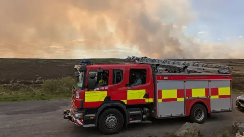Michael Fisher A large fire engine sits in the forefront of the image as a large plume of smoke is in the back.