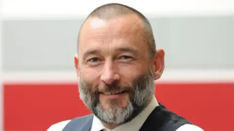 A smiling Peter Hughes looks directly at the camera. He is wearing a waistcoat and white shirt with a red tie. He has a beard and short hair. 