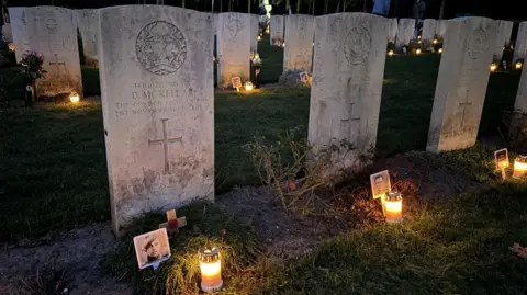 Venray War Cemetery Adoption Graves Foundation Close up image of graves at the British war cemetery in Venray, Netherlands. All the graves have a small bright lantern on the ground in front of them and most have a photograph of the soldier buried there. 