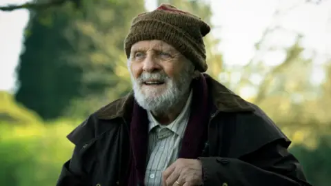 Sir Anthony Hopkins with a grey beard. He wears a dark coat, woollen brown hat and a beige shirt with a blue stripes.
