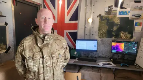 A white man in a green camouflage army outfit. He is standing in a bunker like room that is an immersive training exercise.  