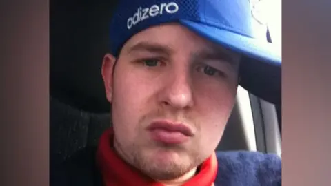 A head and shoulders shot of a man looking at a camera. He wears a blue cap on his head which reads "adizero" on the side. He wears a red and navy top.