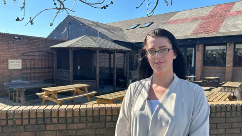 BBC Chanel Walsh standing near a pub garden. There are benches behind her and there is a St. Georges flag on the roof. Behind her there is a shelter.