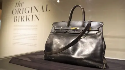 A back leather bag in a display case in front of a display manel saying "The original Birkin"
