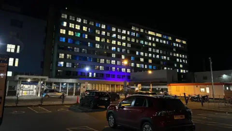 LDRS A car park at night at a hospital. A few cars can be seen in the car park. An "Entrance" sign is at the front of the car park with a large building behind which has dozens of square windows.