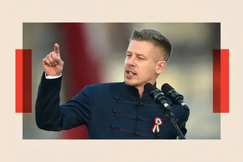 NurPhoto via Getty Images Peter Magyar gesticulates from behind a podium. He wears a high-necked blue suit jacket with brocading on the front, and has a rosette pinned to his chest