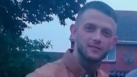 Family Handout A slightly blurred picture of Mr Fowler who is smiling for the camera. He wears a coral coloured shirt and stands in front of a house.