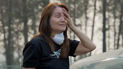 A woman holds her head with her hand. Her hands are closed and she has a face mask loose around her neck.