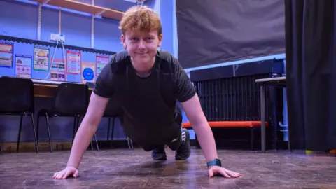 Adam is lying in the push-up position and is smiling at the camera, he is wearing a black t-shirt and black joggers with trainers. 