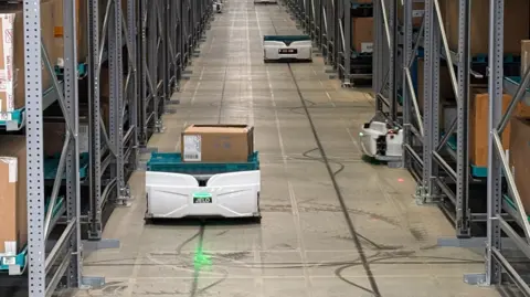 DJ McLaren/BBC A robot carrying a cardboard box along a corridor between rows of tall shelving. The robot is white, with letters "JELO" on its front underneath a green light, whose beam is also reflected on the floor in front. On top of the robot is a green crate with a cardboard box inside. There are several other identical robots behind this one. The floor between the shelves is polished concrete and is light grey in colour, but has black lines up, down and across it, where the robots' wheels have left marks. The shelves are tightly packed, with their metal frames fixed to the floor.