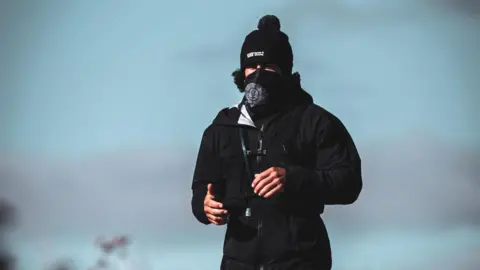 Charlie Probets Mike Humphreys running, wearing black trousers, a black coat, a black scarf over his face and a black bobble hat.
