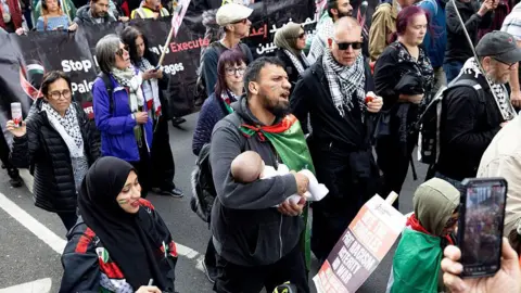 Getty Images About a dozen people are seen in this picture of a portion of the Nakba Anniversary march. A man, wearing a Palestinian flag, can be seen holding a small baby as he walks alongside several other men and women
