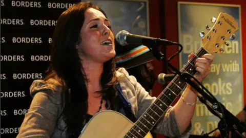PA A girl with dark hair sits at a microphone singing with a wooden guitar on her lap.