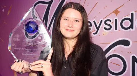 Stephanie Barney has long, dark brown hair and smiles for the cameras while holding her large trophy for winning the Merseyside Woman of the Year award.