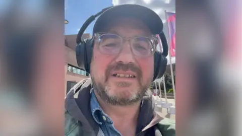 Matthew Carr/YouTube A still from one of Mr Carr's videos. He is standing outside and addressing the camera while wearing headphones and a green raincoat. He has a brown beard