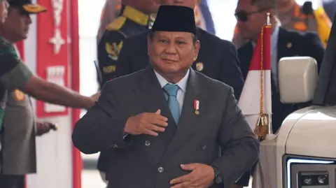 Getty Images Indonésia O presidente da Indonésia, Prabowo Subianto, passa por um cordão de segurança vestindo um terno e com as mãos na jaqueta.
