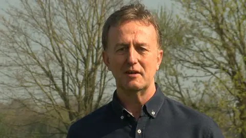 Tim Ward pictured in a field. There are trees behind him. He's wearing a navy oxford shirt and has short dark hair.