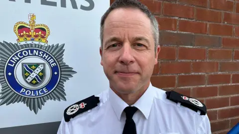Chief Constable Paul Gibson with short dark hair he is wearing a white uniform shirt with epaulettes and is stood in front of a brick wall which has the forces badge on it